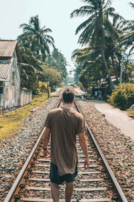man in brown shirt standing on train rail near coconut palms